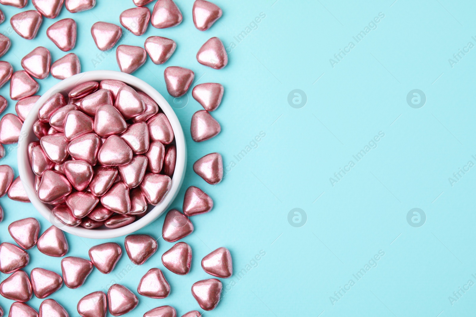 Photo of Bowl and delicious heart shaped candies on light blue background, flat lay. Space for text