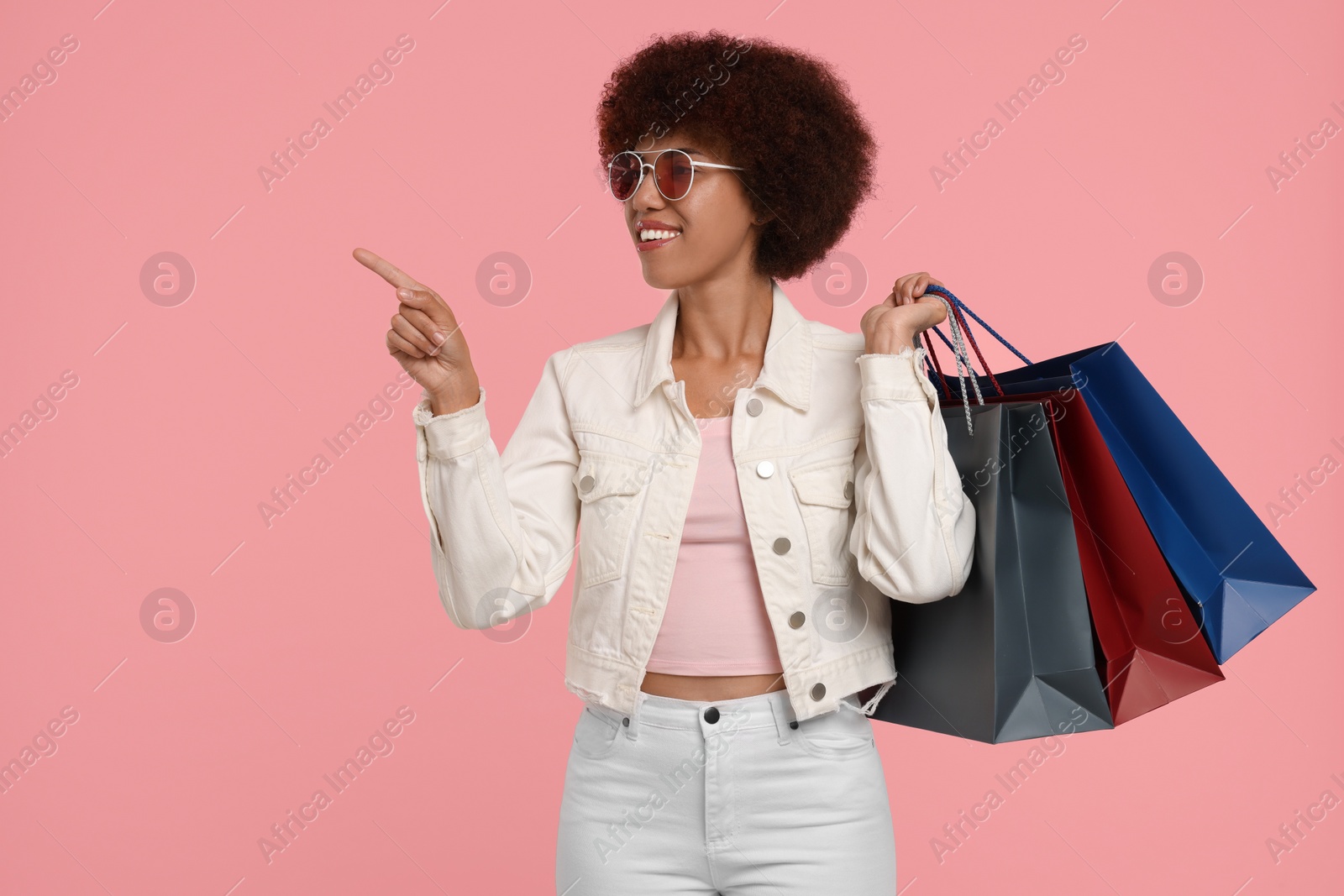 Photo of Happy young woman with shopping bags pointing at something on pink background. Space for text
