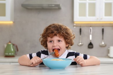 Photo of Cute little boy eating sausage and pasta at table in kitchen