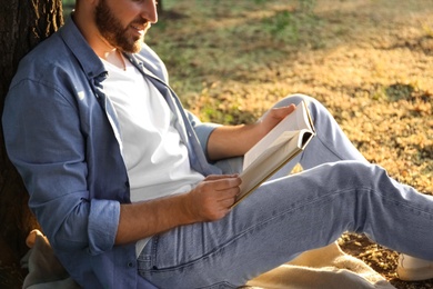 Young man reading book on green grass near tree in park, closeup
