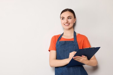 Beautiful young woman in clean denim apron with clipboard on light grey background. Space for text