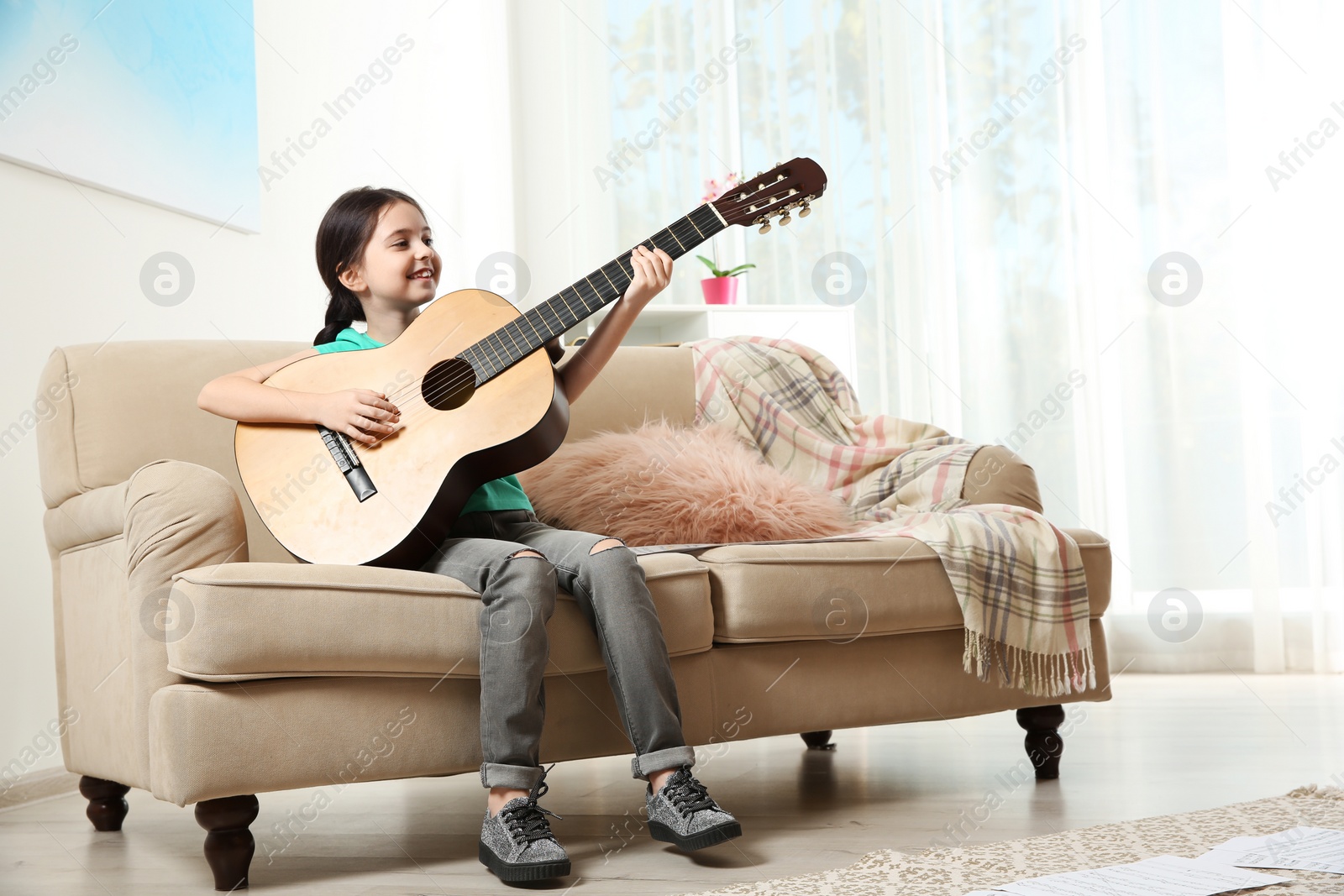 Photo of Cute little girl playing guitar on sofa in room. Space for text