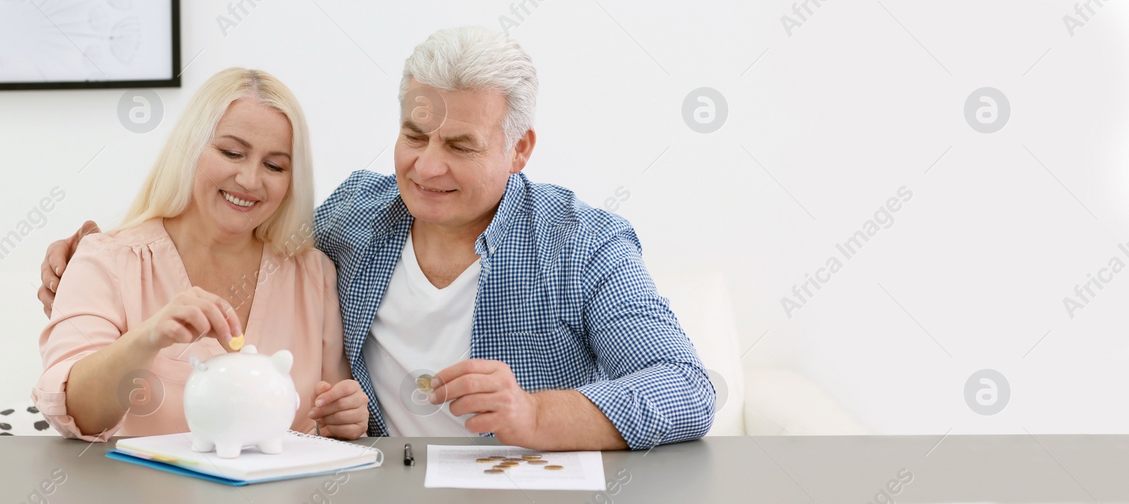 Image of Mature couple putting coin in piggy bank at table, space for text. Banner design