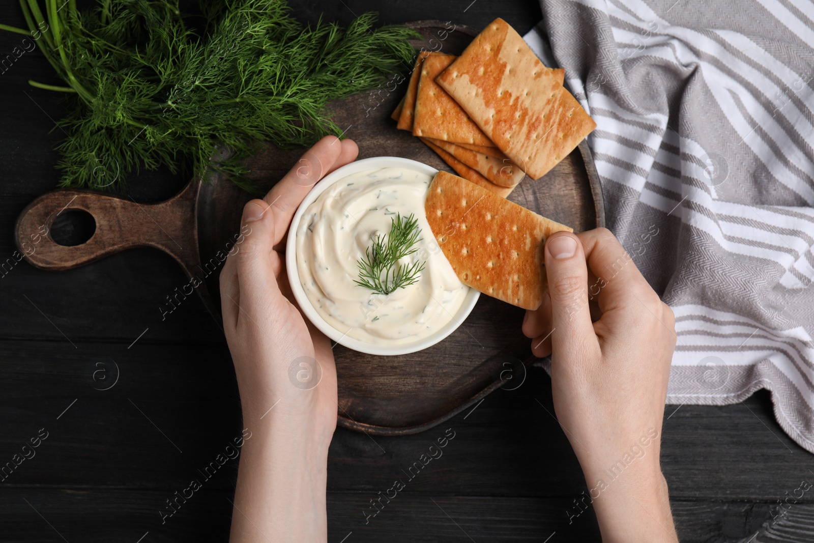 Photo of Woman dipping cracker into tasty creamy dill sauce at black wooden table, top view