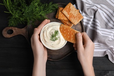 Photo of Woman dipping cracker into tasty creamy dill sauce at black wooden table, top view