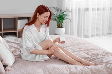 Beautiful young woman applying body cream onto legs in bedroom, space for text