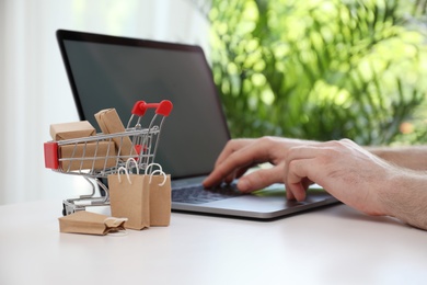 Internet shopping. Small cart with boxes and bags near man using laptop indoors
