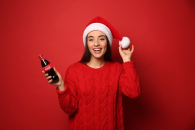 MYKOLAIV, UKRAINE - JANUARY 27, 2021: Young woman in Christmas hat holding bottle of Coca-Cola on red background