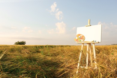 Photo of Wooden easel with blank canvas and painting equipment in field. Space for text