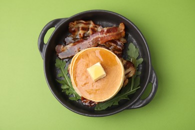 Tasty pancakes with butter, fried bacon and fresh arugula on light green background, top view