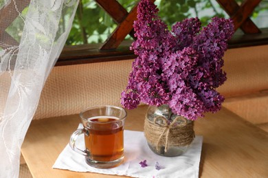 Photo of Bouquet with beautiful lilac flowers and glass cup of tea on wooden table indoors
