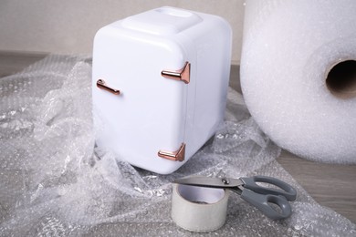 Photo of Cosmetics refrigerator with bubble wrap, scissors and adhesive tape on wooden table