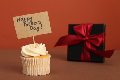 Photo of Happy Father's Day. Tasty cupcake with greetings and gift box on brown table, closeup