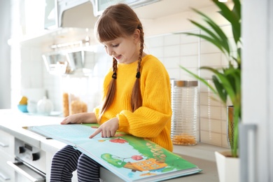 Photo of Cute little girl reading book in kitchen at home