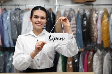 Photo of Dry-cleaning service. Happy worker holding Open sign at counter indoors, space for text
