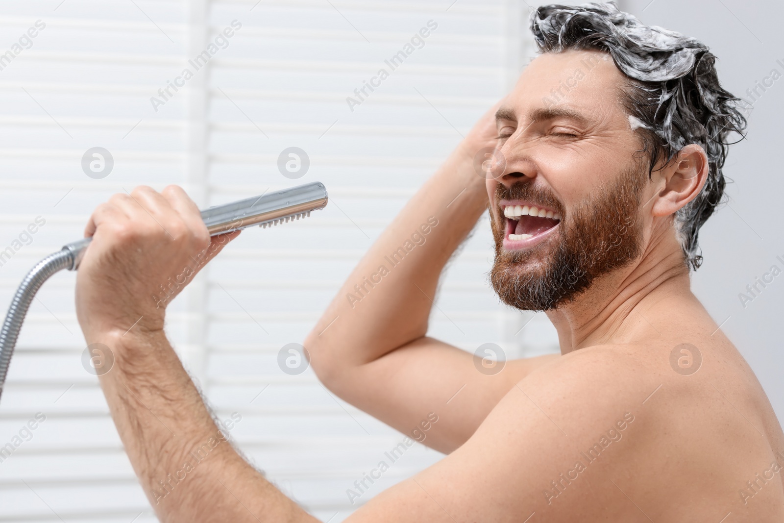 Photo of Happy man with showerhead singing and washing his hair with shampoo indoors