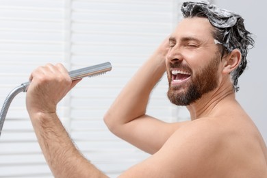 Happy man with showerhead singing and washing his hair with shampoo indoors