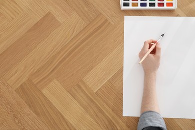 Photo of Man painting with watercolor on blank paper at wooden table, top view. Space for text