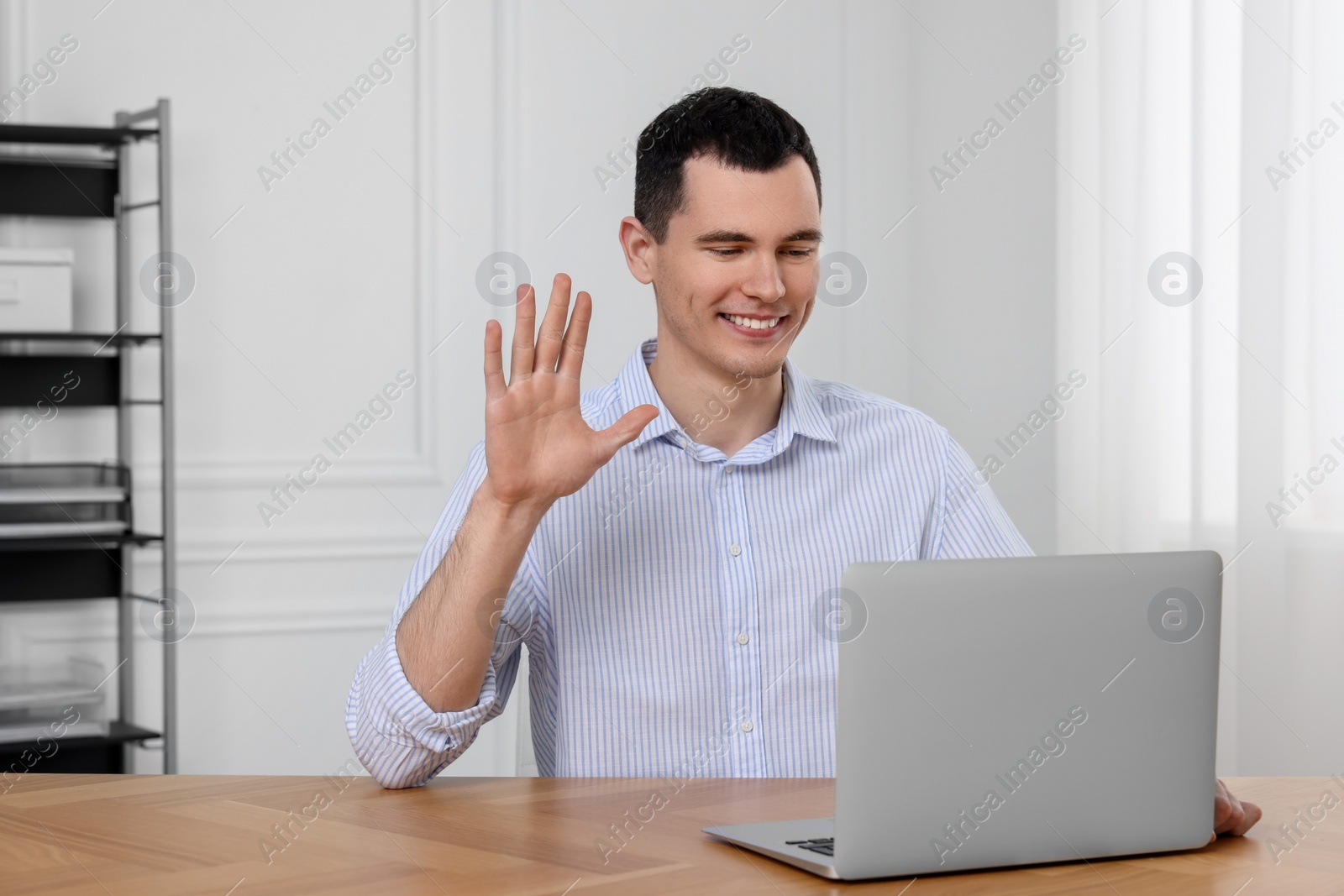 Photo of Man waving hello while having video chat on laptop in office