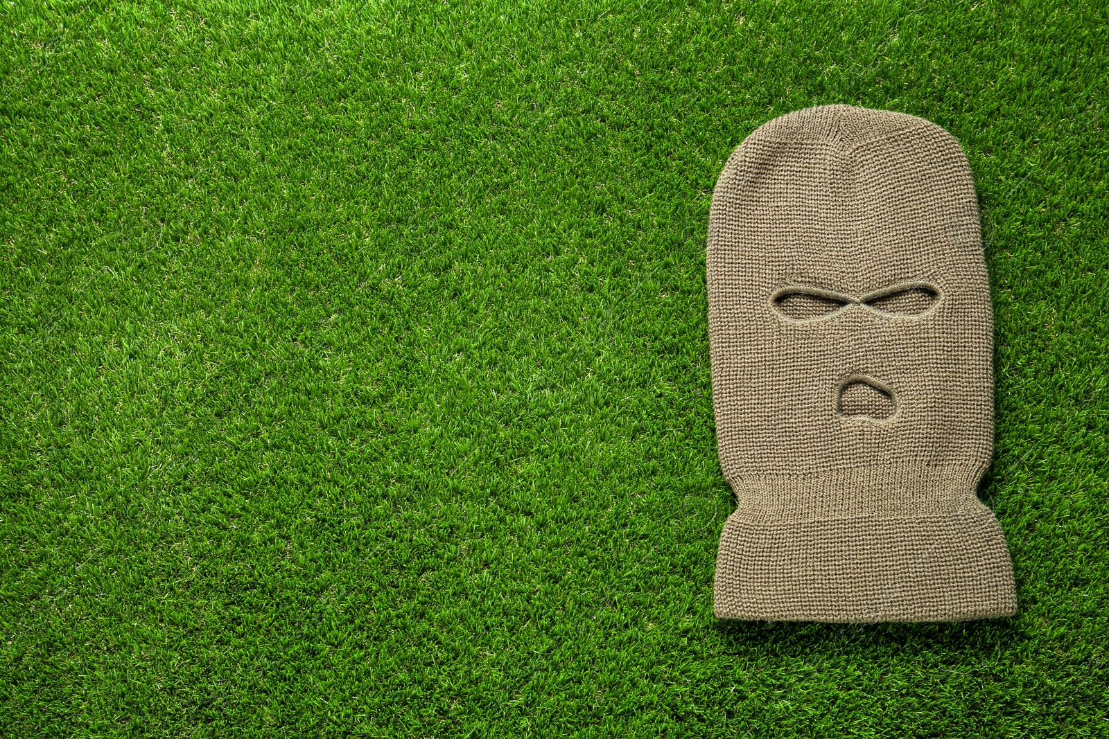 Photo of Beige knitted balaclava on green grass, top view. Space for text