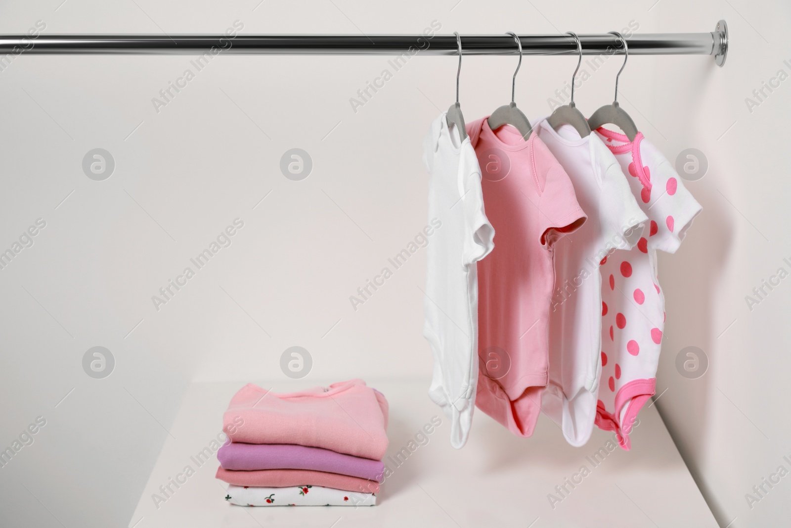 Photo of Hangers with baby bodysuits and stack of clothes near white wall. Space for text