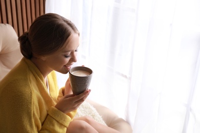 Beautiful young woman drinking coffee near window at home, space for text. Winter atmosphere