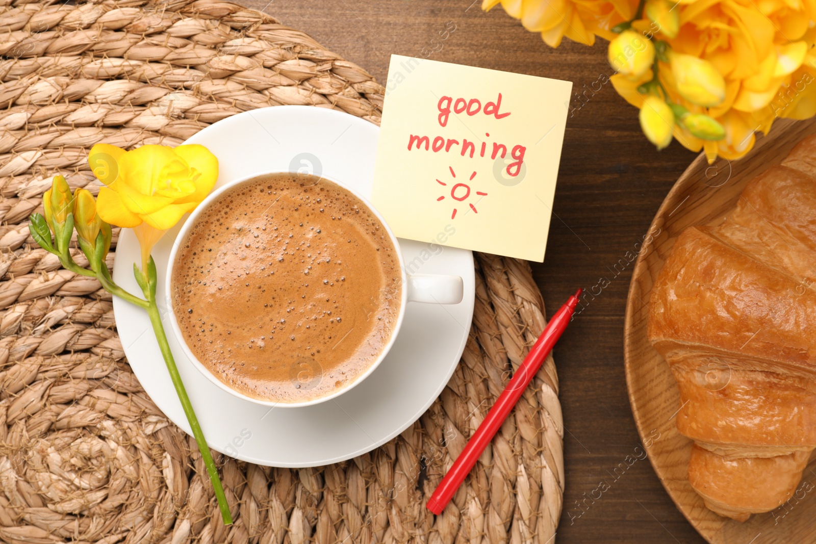 Photo of Cup of aromatic coffee with croissant, beautiful yellow freesias and Good Morning note on wooden table, flat lay