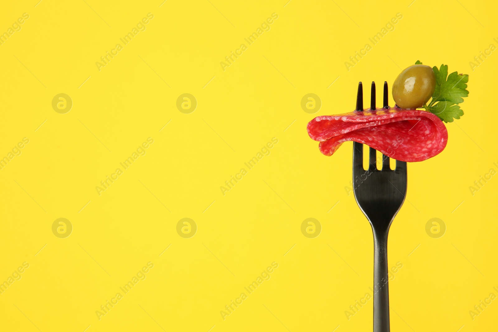 Photo of Fork with tasty slice of salami, olive and parsley on yellow background, space for text