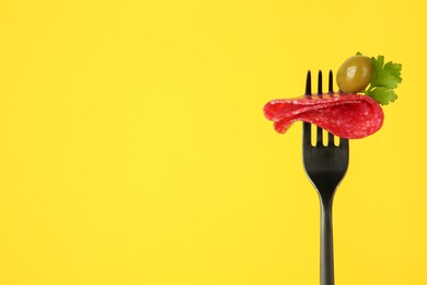 Fork with tasty slice of salami, olive and parsley on yellow background, space for text