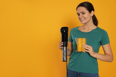 Photo of Beautiful young woman holding sous vide cooker and corn in vacuum pack on orange background. Space for text