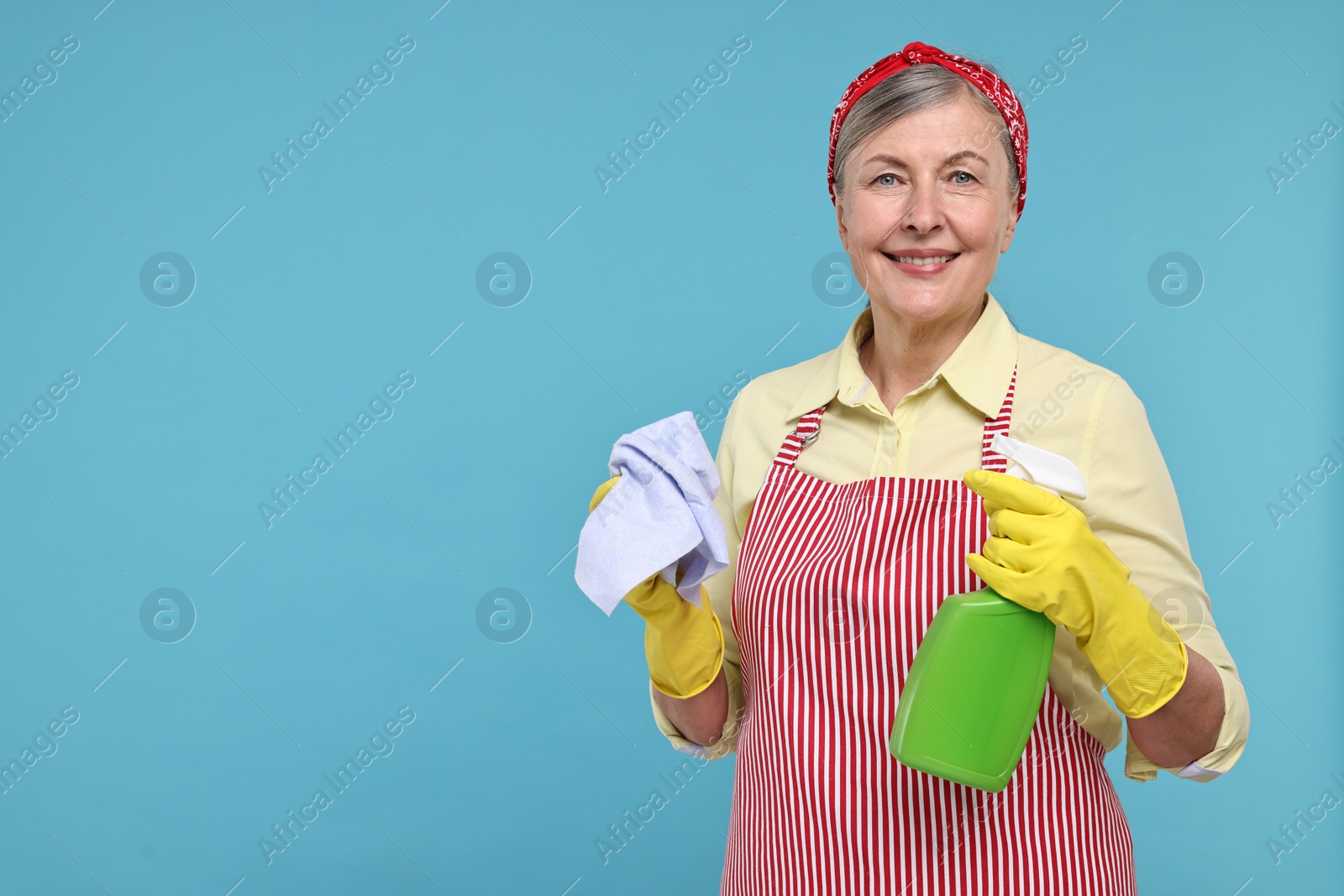 Photo of Happy housewife with spray bottle and rag on light blue background, space for text