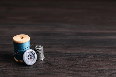 Photo of Spool of light blue thread with needle, button and thimble on wooden table, space for text. Sewing accessories