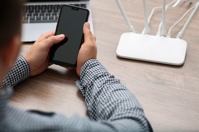 Photo of Man with smartphone and laptop connecting to internet via Wi-Fi router at wooden table, closeup