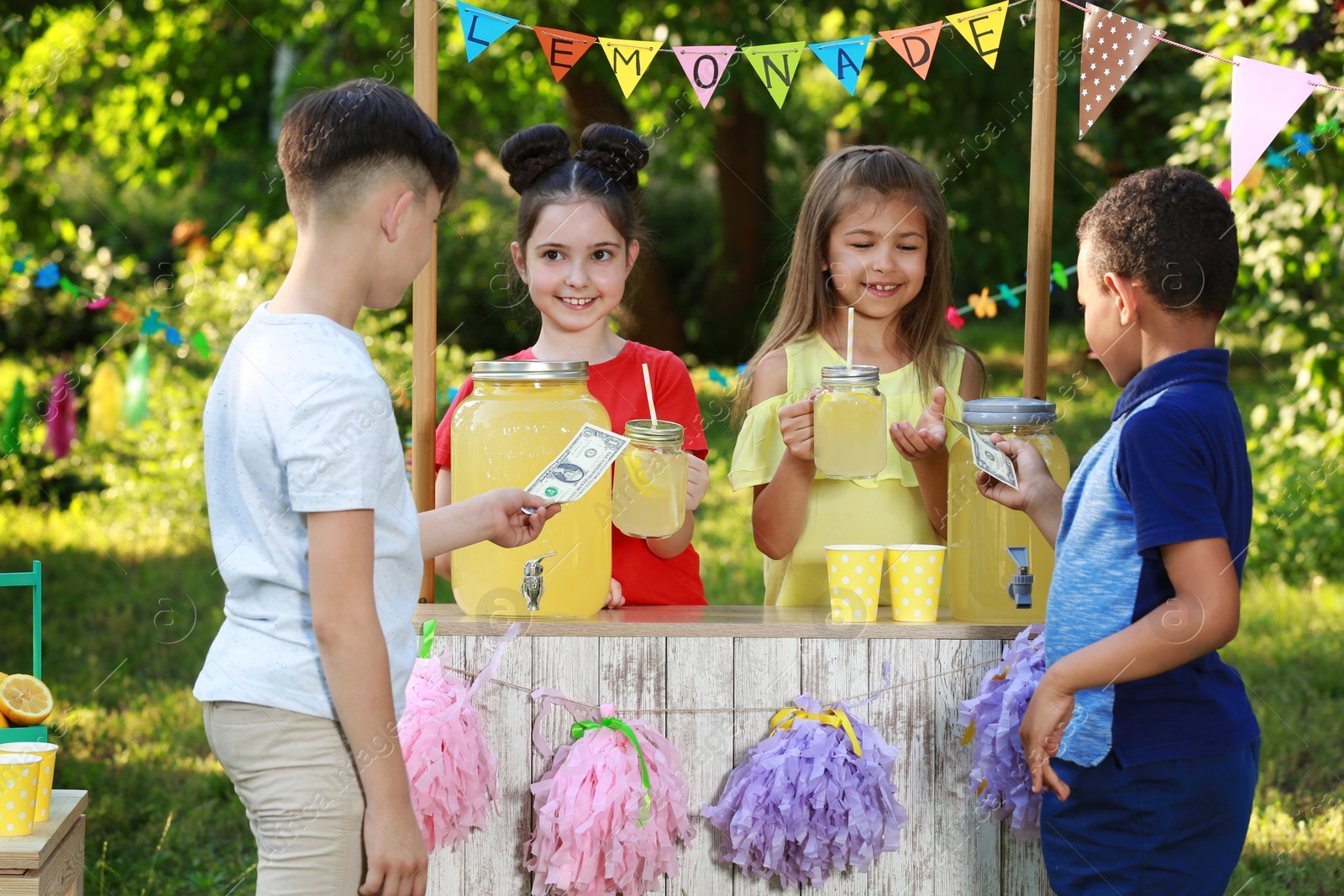Photo of Cute little girls selling natural lemonade to boys in park. Summer refreshing drink