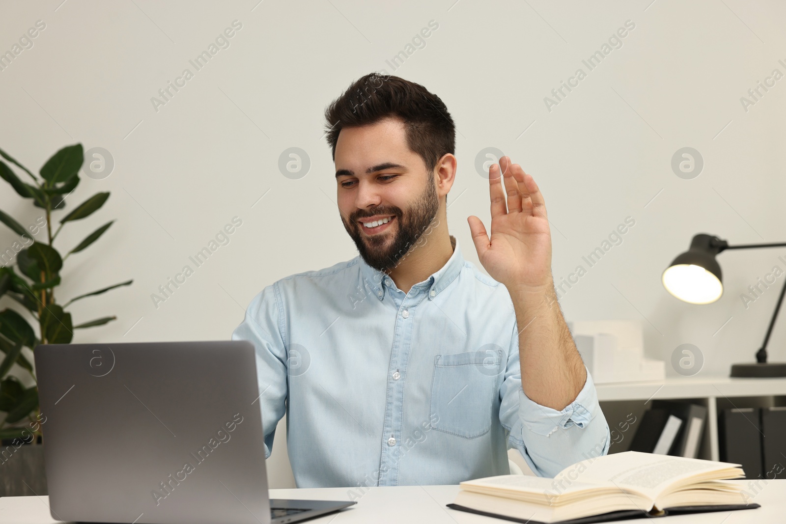 Photo of Young man waving hello during video chat via laptop at table indoors