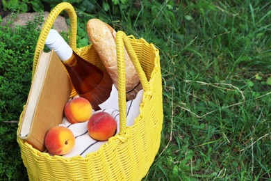 Yellow wicker bag with book, peaches, baguette and wine on green grass outdoors