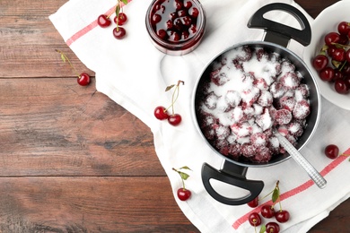 Flat lay composition of pot with cherries and sugar on wooden table, space for text. Making delicious jam