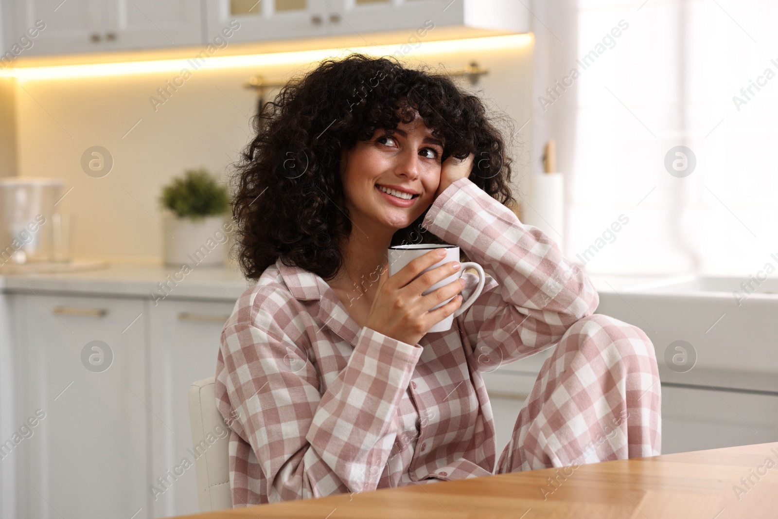 Photo of Beautiful young woman in stylish pyjama with cup of drink at wooden table in kitchen