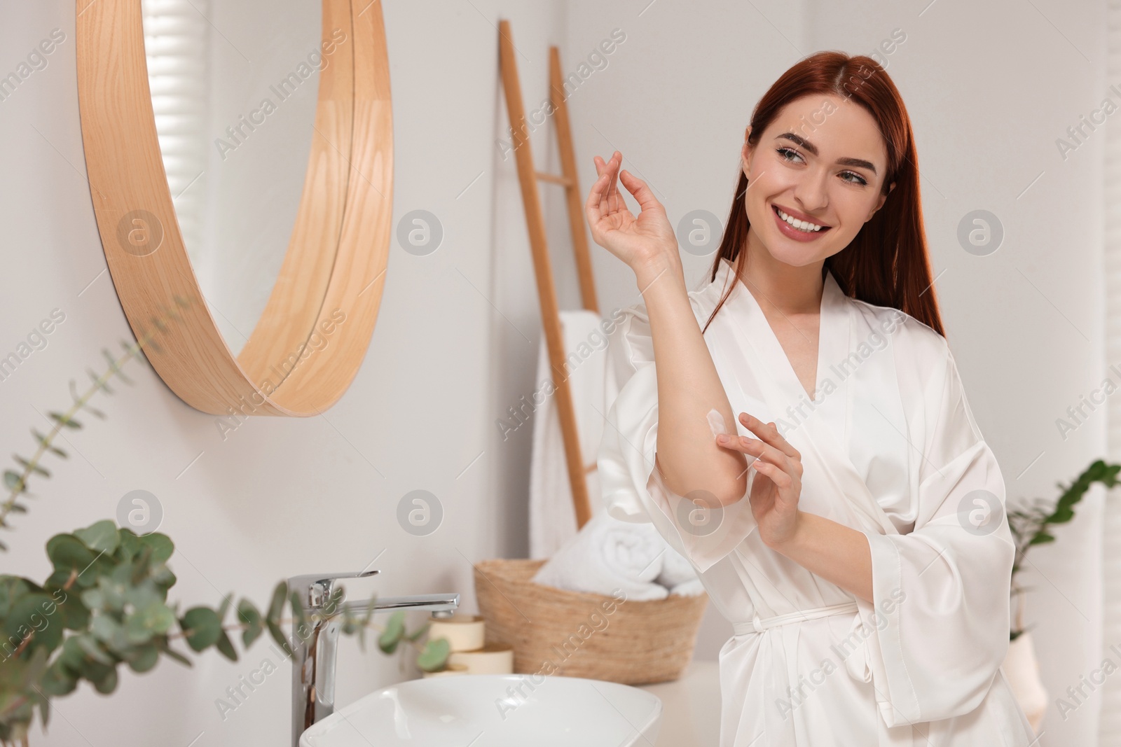 Photo of Beautiful young woman applying body cream onto elbow in bathroom, space for text