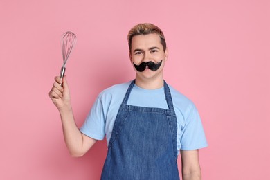 Photo of Portrait of happy confectioner with funny artificial moustache holding whisk on pink background