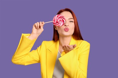 Stylish redhead woman covering eye with lollipop and blowing kiss on purple background