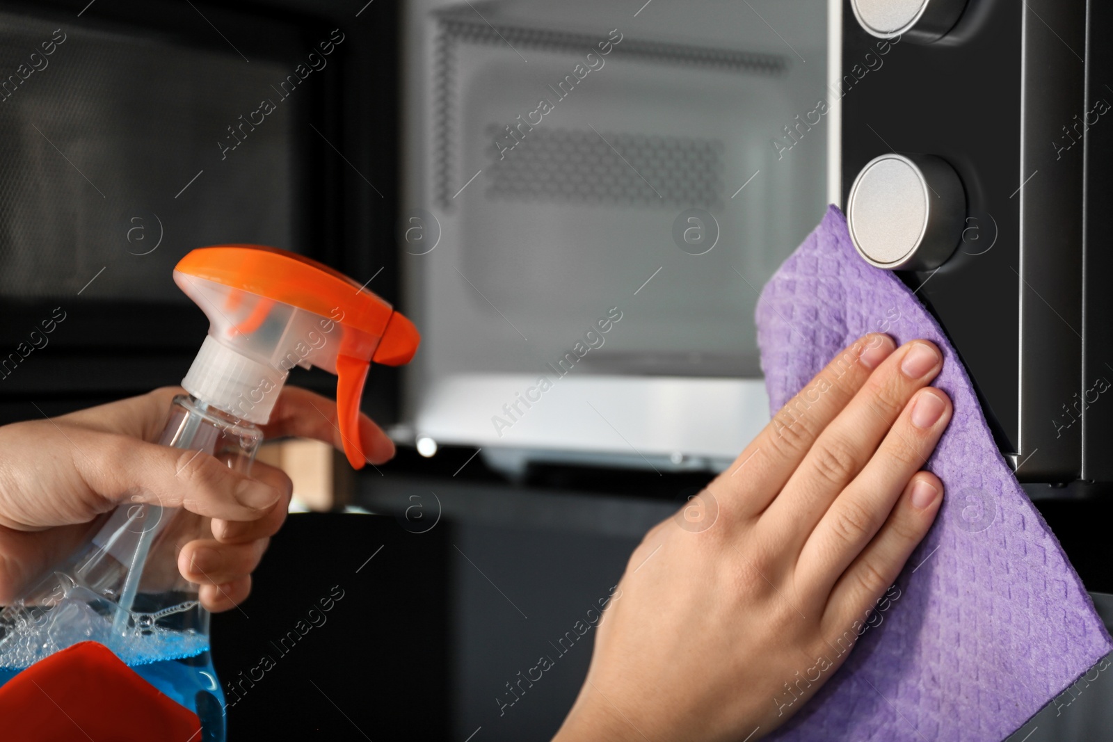 Photo of Woman cleaning microwave oven with rag and detergent in kitchen, closeup