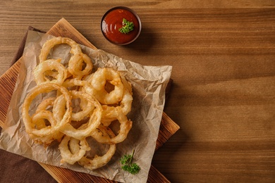 Flat lay composition with homemade crispy onion rings and tomato sauce on wooden background. Space for text