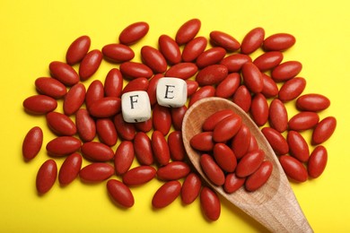 Photo of Wooden cubes with letters FE, spoon and red pills on yellow background, flat lay. Anemia treatment
