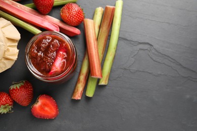 Photo of Jar of tasty rhubarb jam, fresh stems and strawberries on dark textured table, flat lay. Space for text