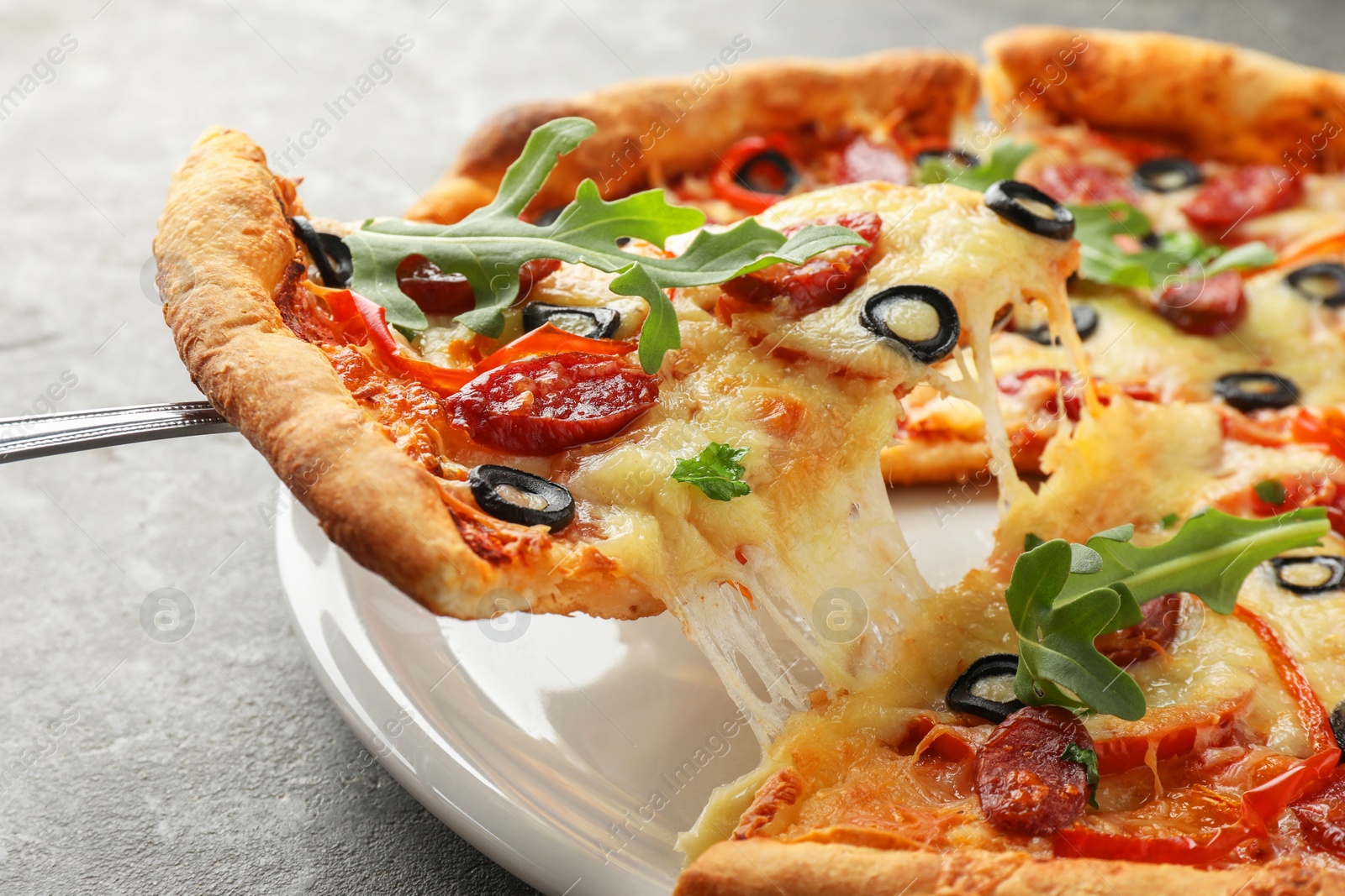 Photo of Taking piece of tasty pizza with smoked sausages, olives, pepper and parsley at gray table, closeup