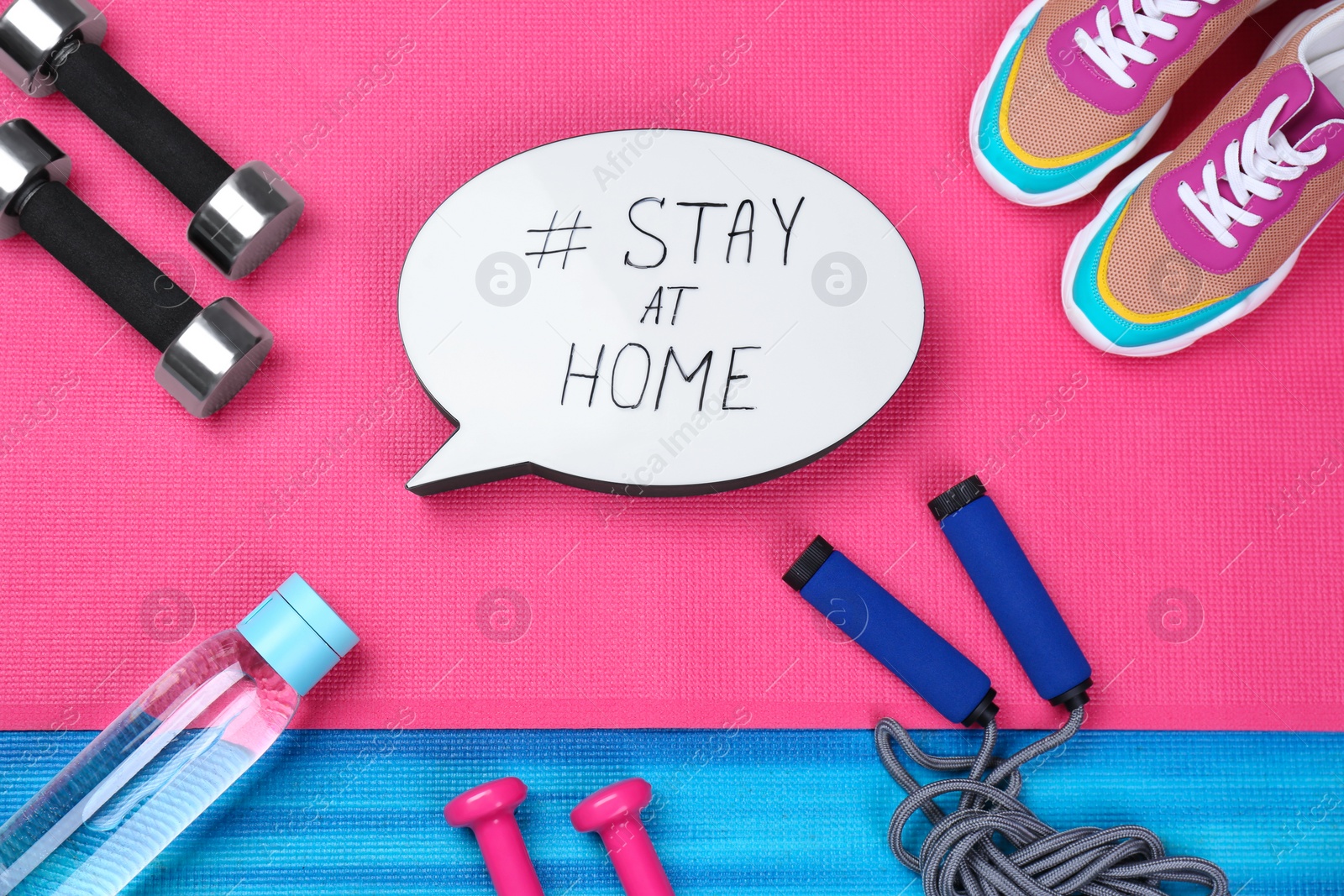 Photo of Sport equipment and speech bubble with hashtag STAY AT HOME on colorful yoga mats, flat lay. Message to promote self-isolation during COVID‑19 pandemic