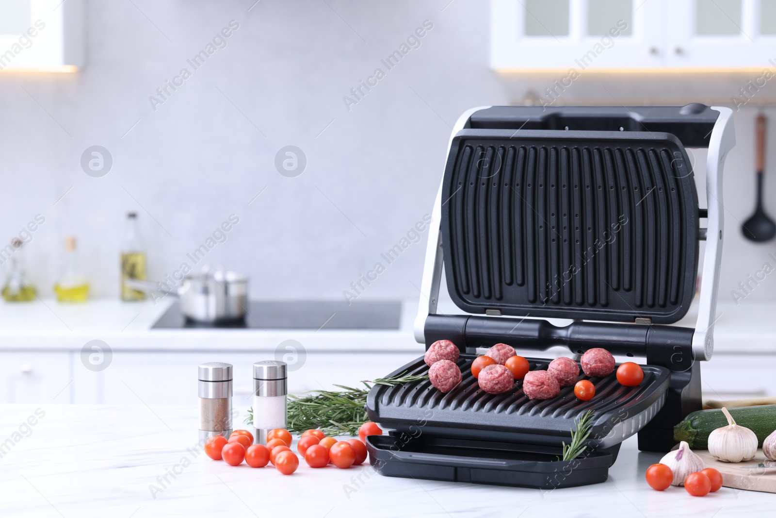 Photo of Electric grill with meatballs, tomatoes, spices and vegetables on white marble table in kitchen. Space for text