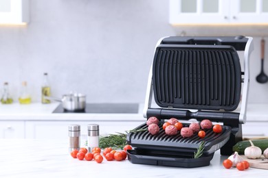 Photo of Electric grill with meatballs, tomatoes, spices and vegetables on white marble table in kitchen. Space for text