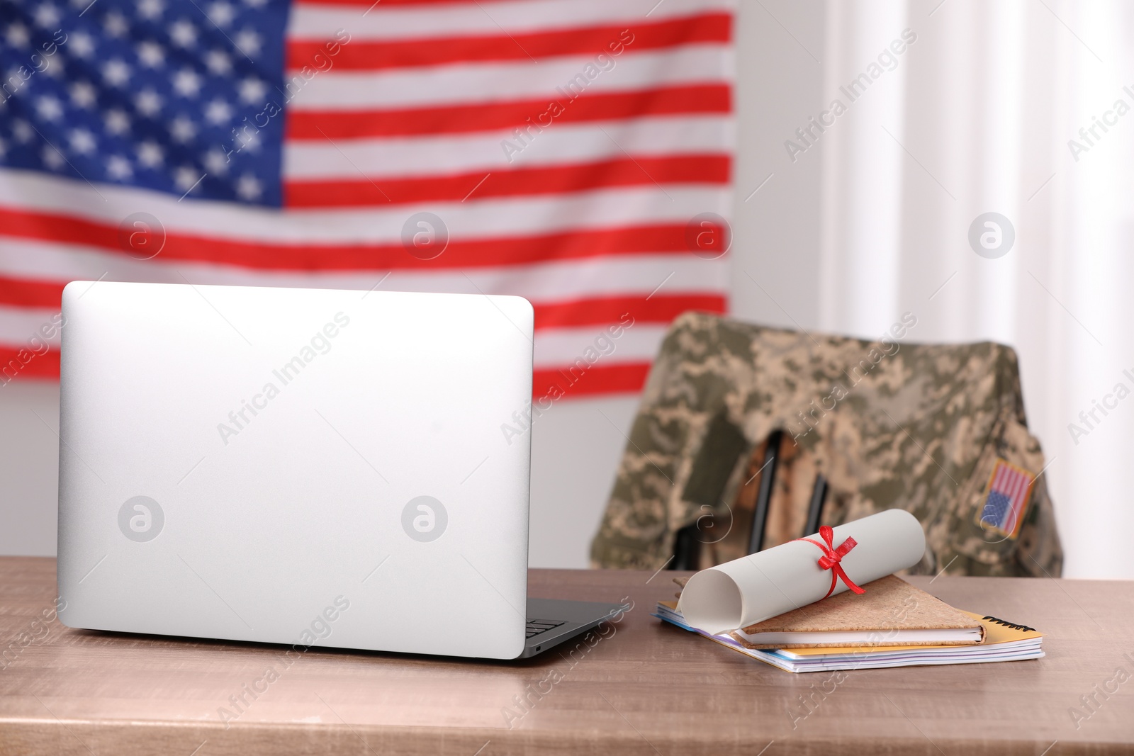 Photo of Laptop, notebooks and diploma on wooden table, chair with soldier uniform against flag of United States indoors. Military education